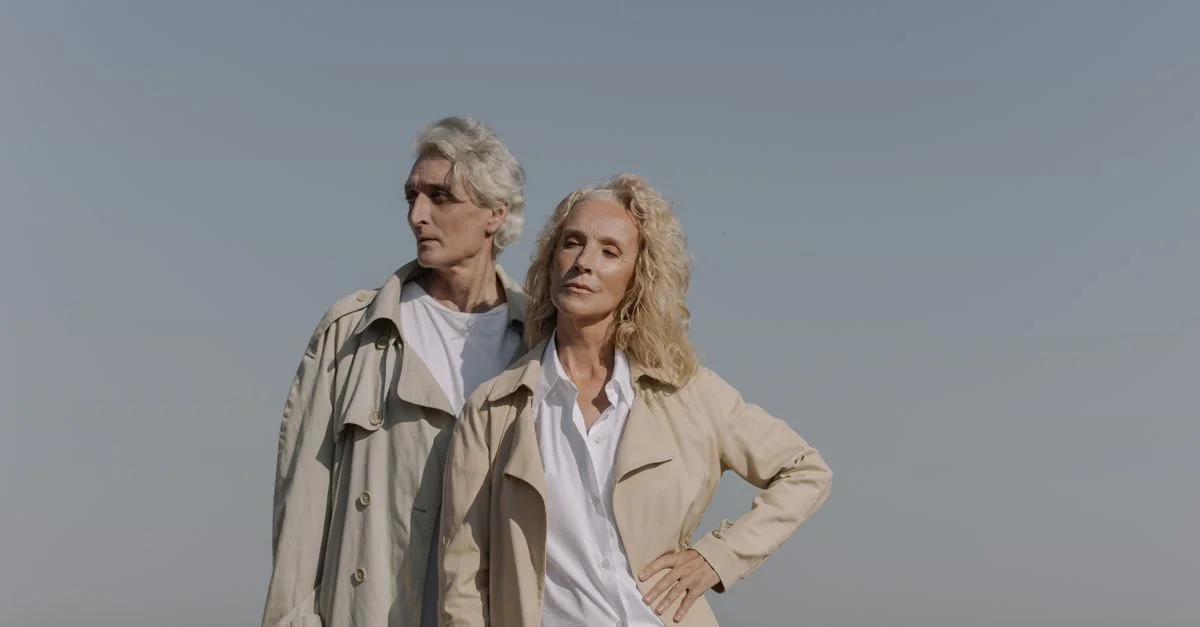 Stylé couple senior posant à la plage, sous un ciel bleu clair, portant des trenchs.