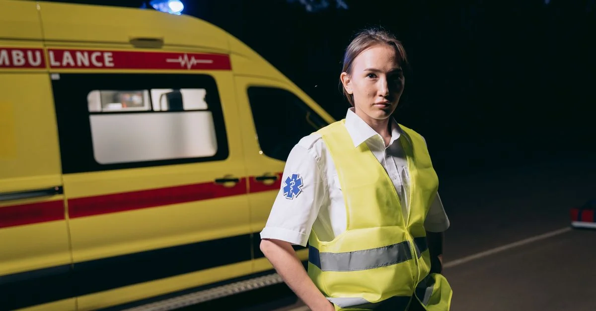 Paramédic femme en gilet réfléchissant à côté de l'ambulance dans la rue de la ville la nuit.