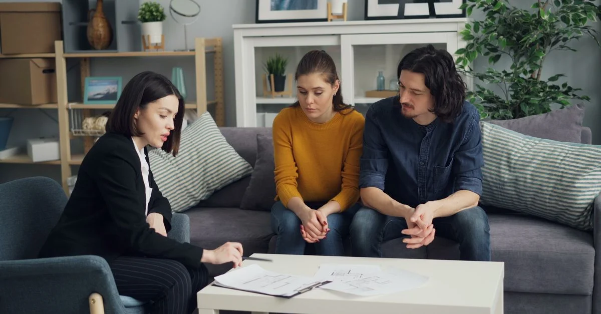 Couple discutant de propriété avec un agent immobilier dans un cadre de salon confortable, examinant des documents.